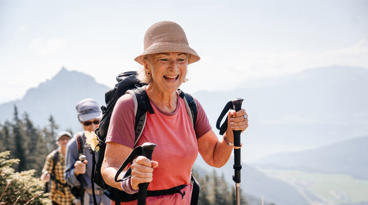 Eine ältere Frau mit Stöcken und Sonnenhut führt glücklich eine Wandergruppe an. | © DAV / Marisa Koch